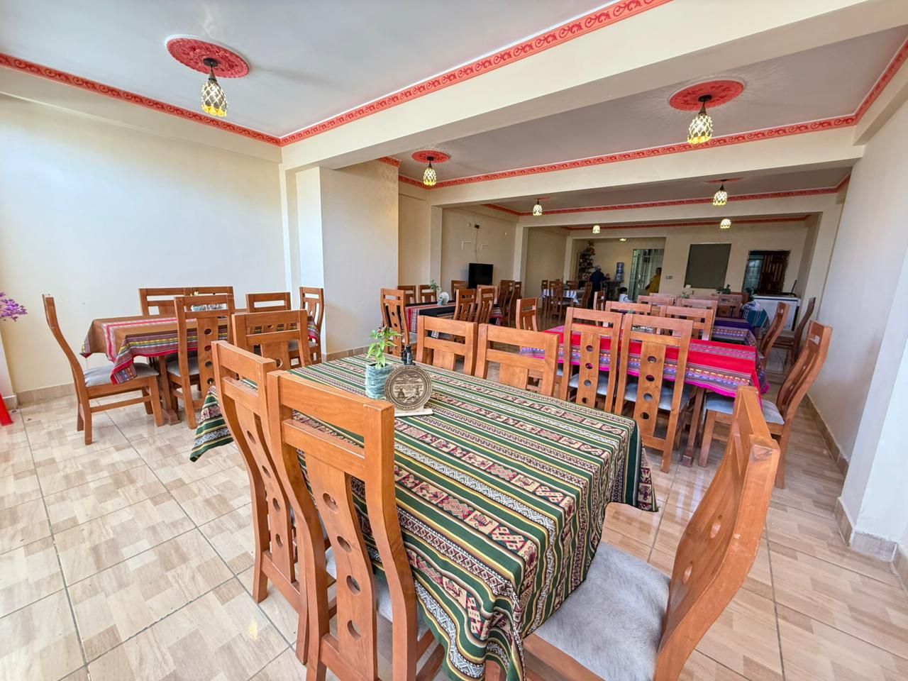 A dining area with multiple wooden tables and chairs, a patterned tablecloth, and decorative ceiling lights.