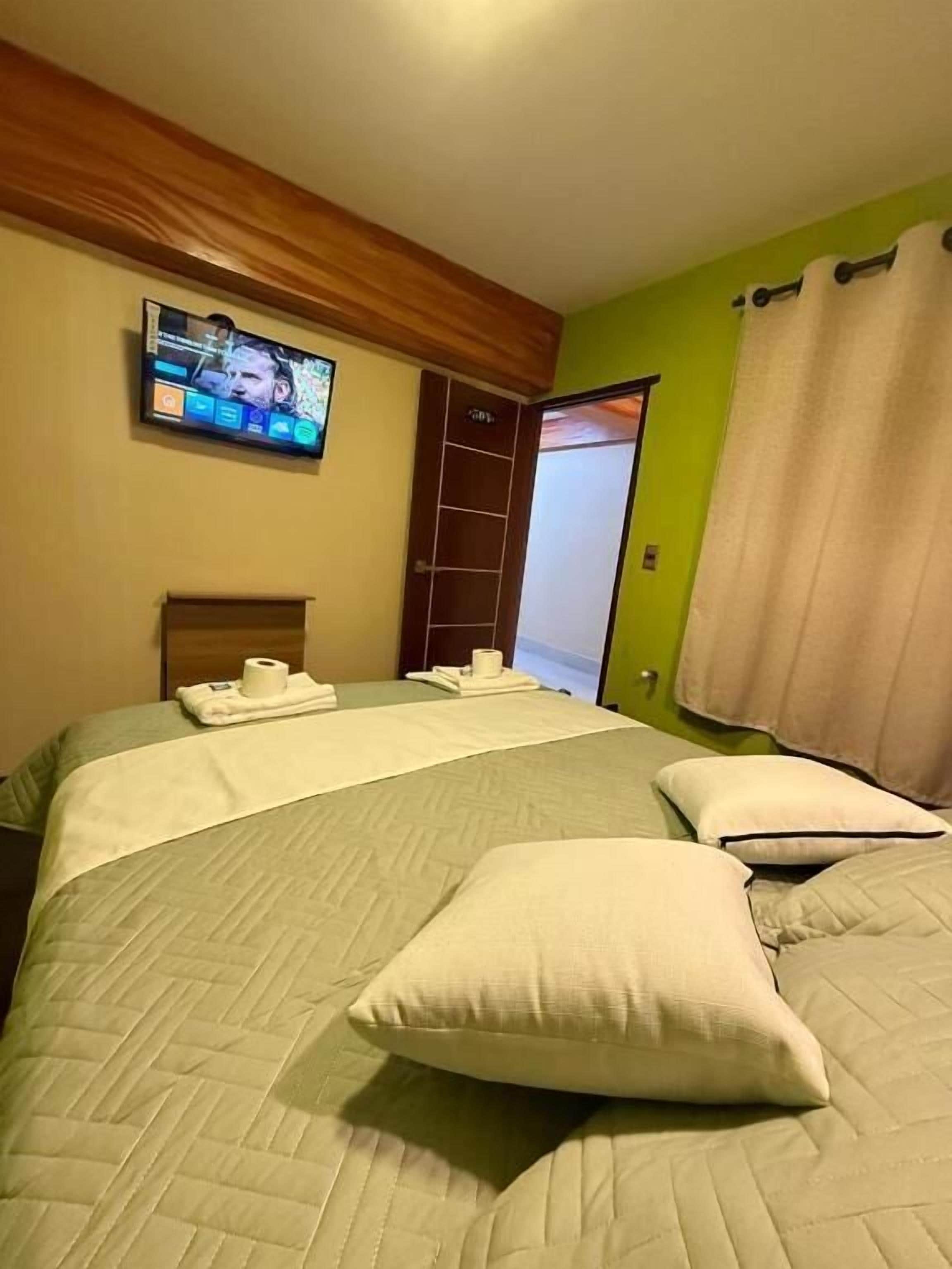 Habitación de hotel con cama, televisor montado en la pared, una puerta y una cortina.
