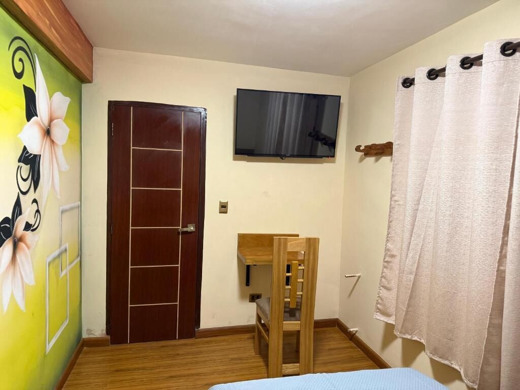 Una habitación con puerta de madera, un televisor montado en la pared, una mesita de madera y una cama con colcha azul.