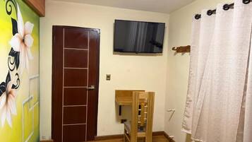 Una habitación con puerta de madera, un televisor montado en la pared, una mesita de madera y una cama con colcha azul.