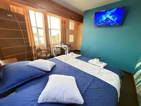 A hotel room with a bed, pillows, a TV, a small table with a chair, and a window with curtains.