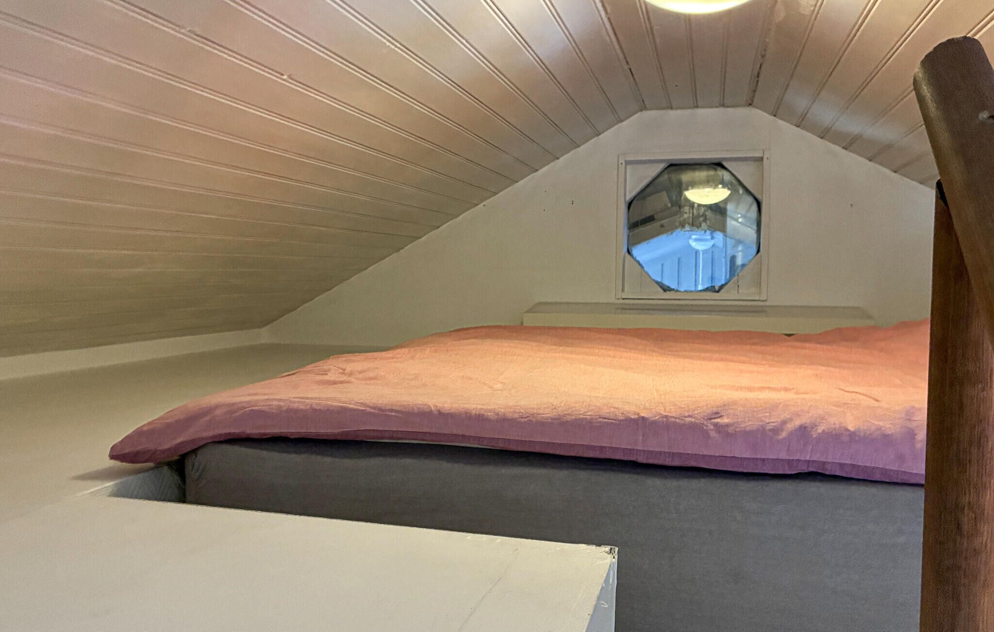 A small attic bedroom with a sloped ceiling, a single window, a bed with a pink mattress, and a wooden headboard.
