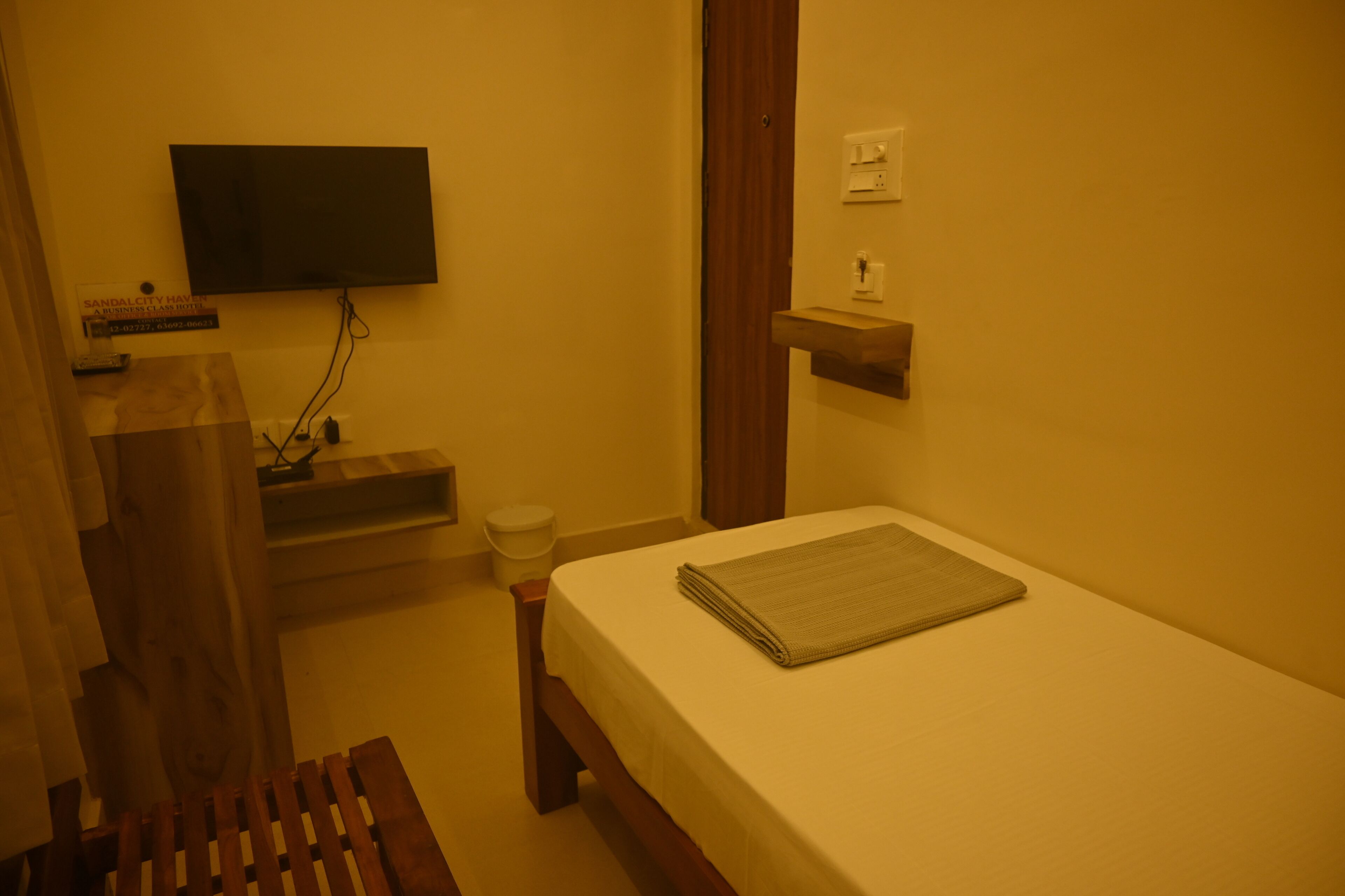 A small room with a bed, a television mounted on the wall, a wooden bench, and a small shelf.