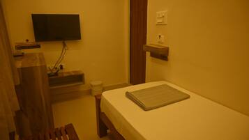 A small room with a bed, a television mounted on the wall, a wooden bench, and a small shelf.