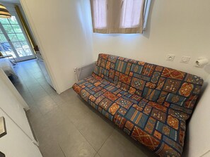 A room with a patterned sofa, a radiator, a window with curtains, and a door leading to another room. - APPARTEMENT SAINT CLAIR (LE LAVANDOU)
