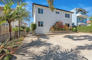 Ein modernes, zweistöckiges Wohnhaus mit weißer Fassade, einem Balkon im oberen Geschoss und einem gepflegten Garten mit Palmen und blühenden Pflanzen.