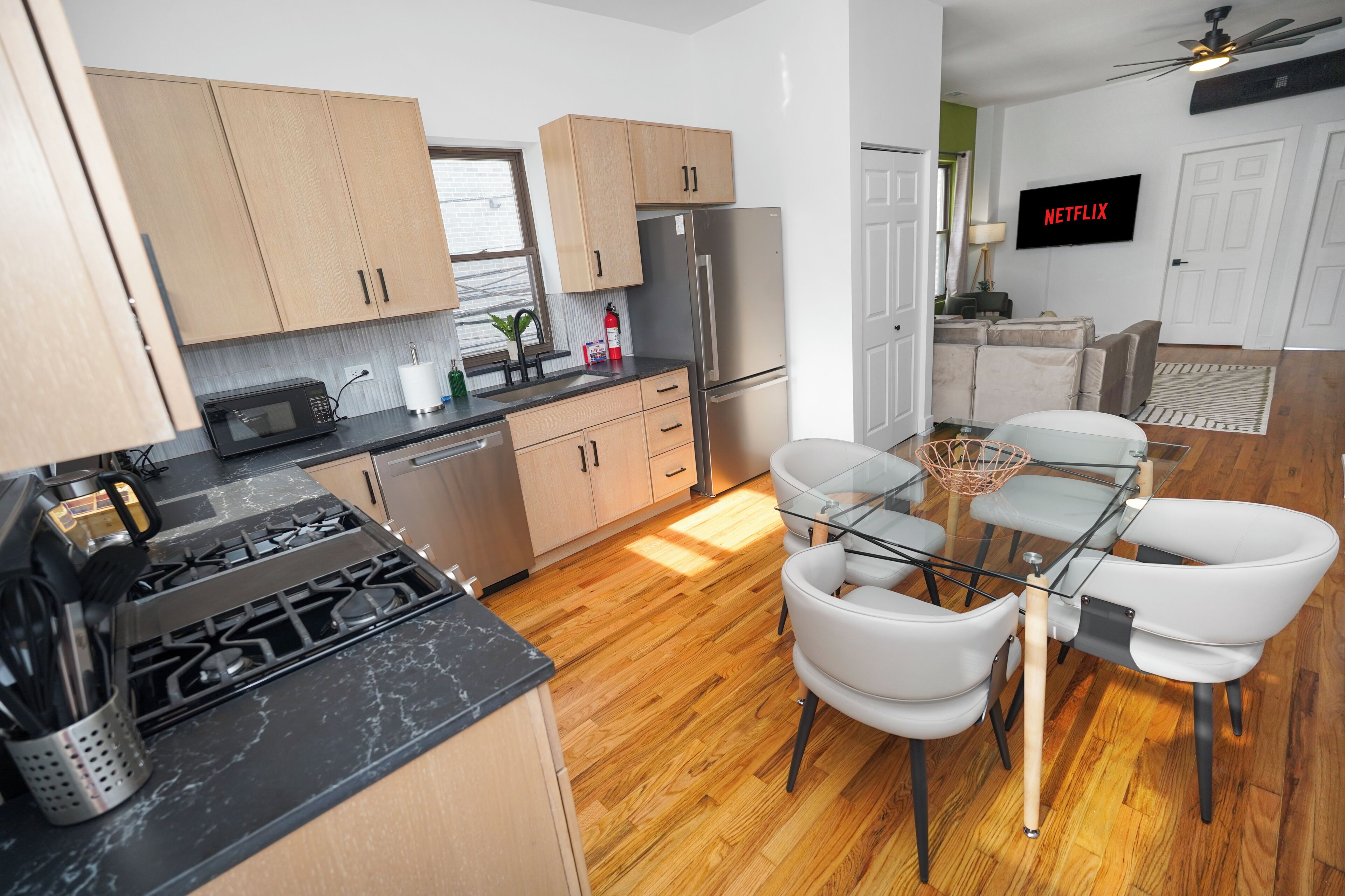 Une cuisine moderne avec des meubles en bois, un réfrigérateur en acier inoxydable, une table à manger en verre entourée de chaises blanches, et un téléviseur affichant le logo « NETFLIX » au mur.