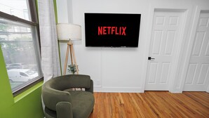 Ein Zimmer mit grüner Wand, Holzboden, einem grünen Sessel, einer Stehlampe und einem Fernseher, der 'NETFLIX' anzeigt.