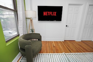 Une pièce avec un mur vert, un plancher en bois, un fauteuil vert, un lampadaire et un téléviseur affichant le logo « NETFLIX ».