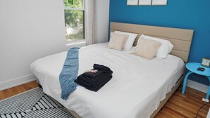 Ein ordentlich bezogenes Bett mit einem blauen Handtuch und einem gefalteten schwarzen Handtuch, ein kleiner runder blauer Tisch mit einer Uhr und ein Fenster mit Blick auf Grünflächen.