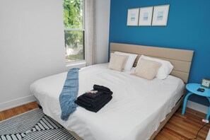 Un lit bien fait, avec une serviette bleue et une serviette noire pliée, une petite table ronde bleue sur laquelle se trouve une horloge, et une fenêtre donnant sur un paysage verdoyant.