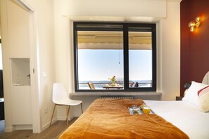 Ein Hotelzimmer mit großem Fenster, einem Bett mit brauner Decke, einem weißen Stuhl und einem Tisch mit Frühstücksgebäck.