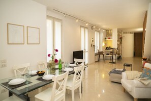 A modern living room with a glass dining table, white chairs, a sofa, and a TV. - Corte Cavour in the City Center (Gallarate)