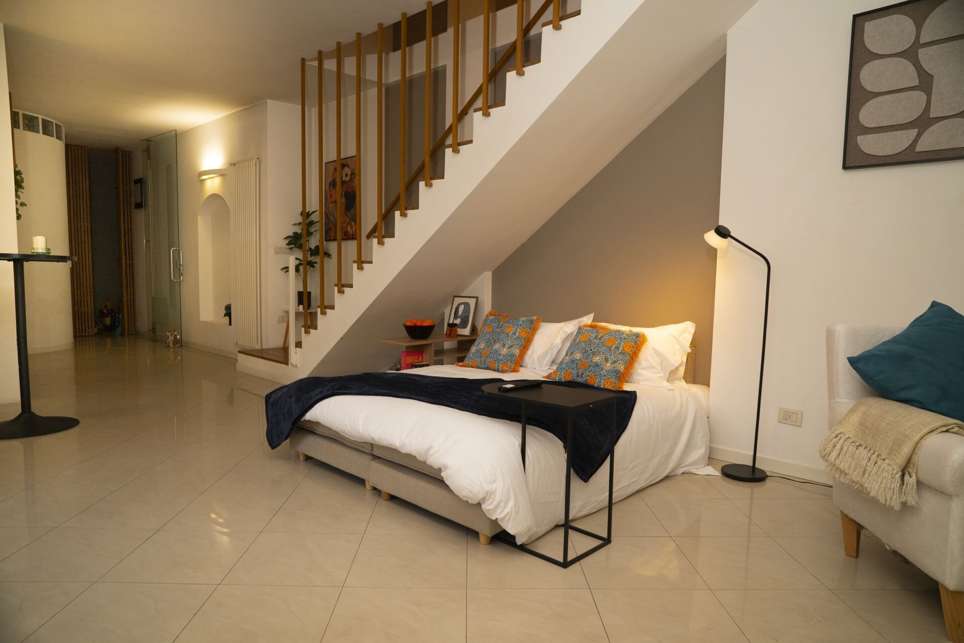 A bedroom with a bed, a nightstand, a lamp, a sofa, a staircase, and a door leading to another room.