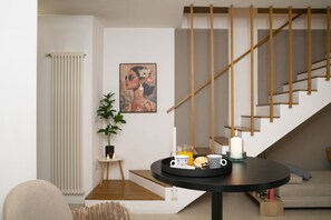 Un salon moderne avec une table ronde noire, un escalier avec des rampes en bois, un radiateur, une plante en pot et une œuvre d’art encadrée accrochée au mur.