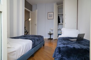 Une chambre avec deux lits, un ordinateur portable posé sur l’un des lits, une petite table avec une horloge et des fleurs, et une armoire remplie de vêtements.