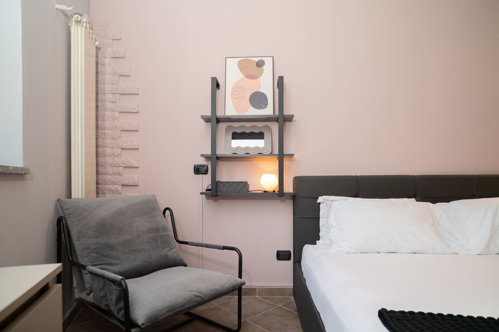 Una camera da letto con un letto, una sedia, uno scaffale con oggetti decorativi e un termosifone.