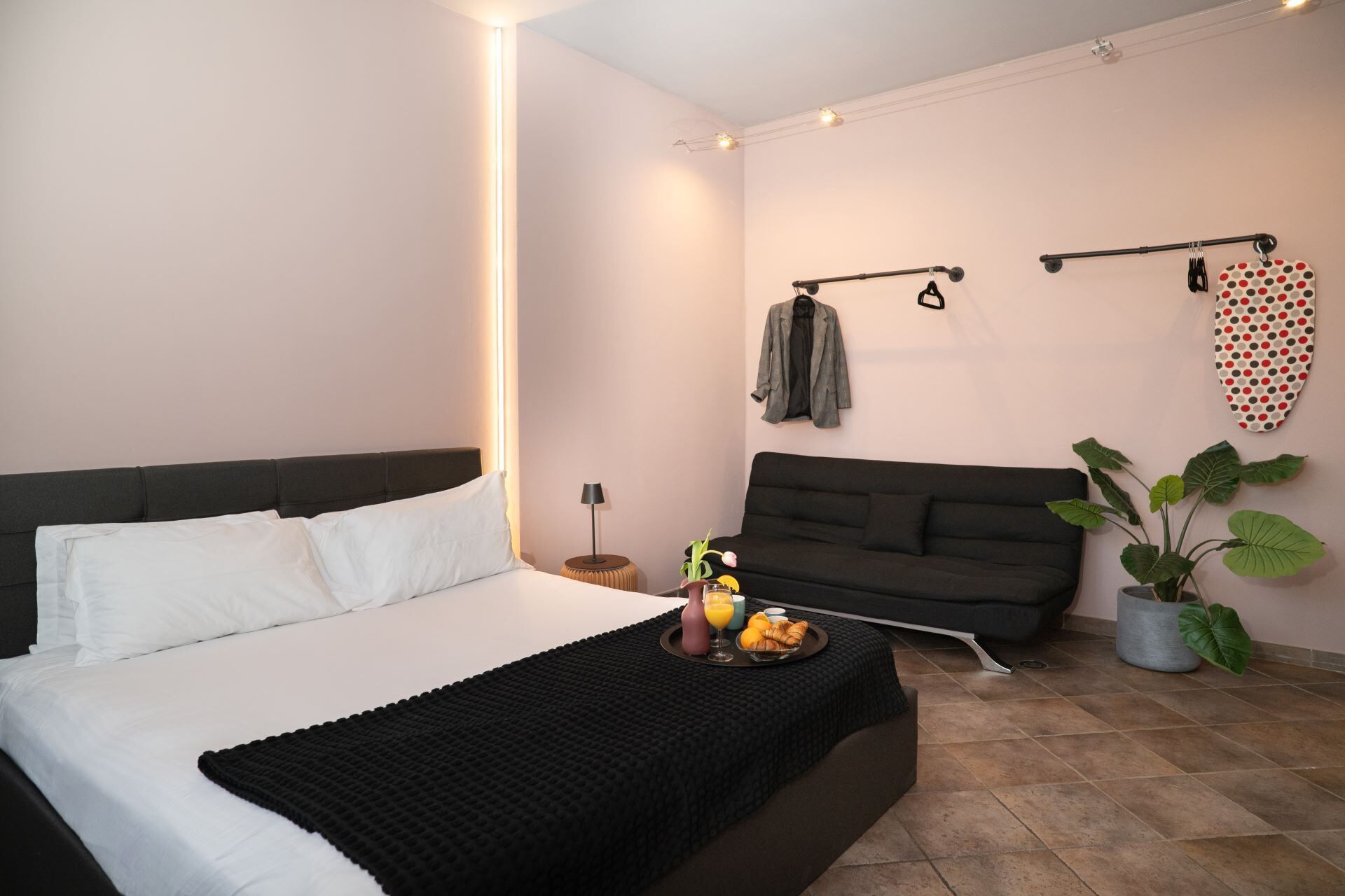 A modern hotel room with a bed, a sofa, a small table with breakfast, a coat rack, and a potted plant.