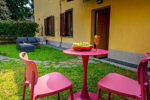 Een geel gebouw met een klein terras, met een roze tafel en stoelen, een schaal fruit en een glas drinken.