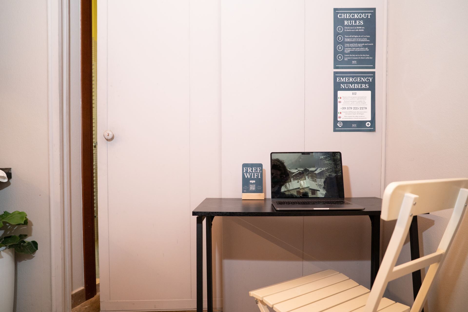 Ett rum med ett skrivbord, en bärbar dator och en stol. På väggen finns anslag med information om utcheckningsregler och telefonnummer för nödsituationer.