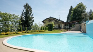 A swimming pool with a curved edge, surrounded by a concrete deck. In the background, there is a building with a curved roof and a staircase leading to a covered area. - Da Gregorio: The Lake View Belvedere with Pools (Gignese)