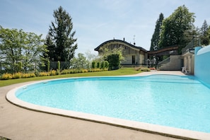 A swimming pool with a curved edge, surrounded by a concrete deck. In the background, there is a building with a curved roof and a staircase leading to a covered area. - Da Gregorio: The Lake View Belvedere with Pools (Gignese)