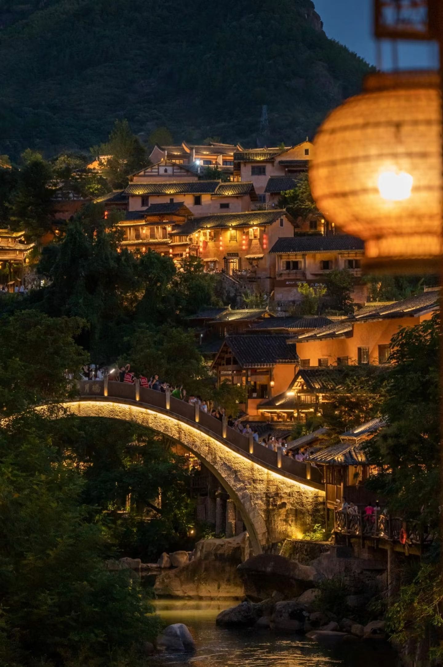 A traditional Chinese village with a stone bridge over a river, surrounded by lush greenery and traditional buildings.
