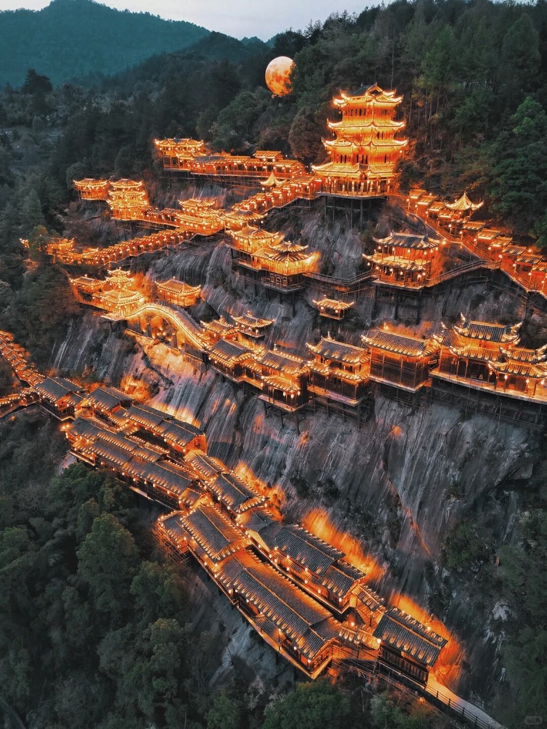A traditional multi-tiered pagoda-style building complex with multiple levels and a central tower, surrounded by steep cliffs and dense forest.