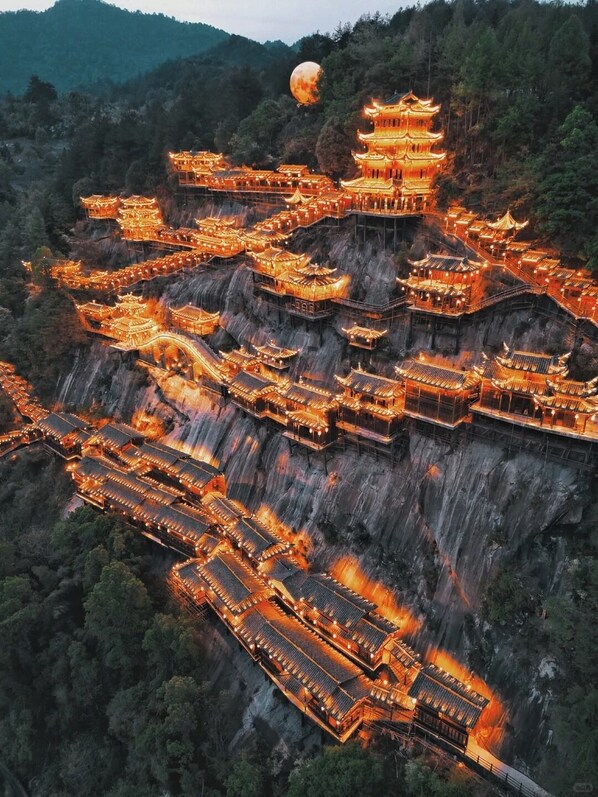 Un complejo arquitectónico tradicional de estilo pagoda, con varios niveles y una torre central, rodeado de acantilados escarpados y un bosque frondoso.