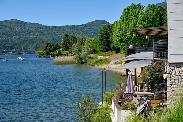A lakeside view with boats, a stone building, outdoor seating, and a mountain backdrop. - Henriette Apt Terrace, Lake Access (Angera)