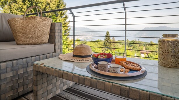 A balcony with a view of mountains, a wicker basket, a straw hat, a bowl of strawberries, a coffee cup, and a glass of orange juice. - LAKESCAPE apartment with terrace, view and pools (Gignese)