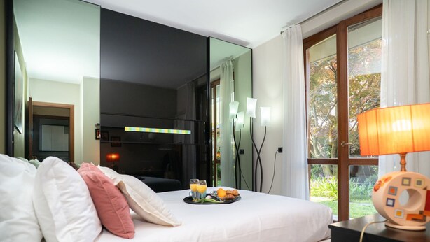 Ein Hotelzimmer mit einem Bett, einem Nachttisch mit Lampe, einem Frühstücksbrettchen und einem großen Fenster mit Blick auf Bäume.