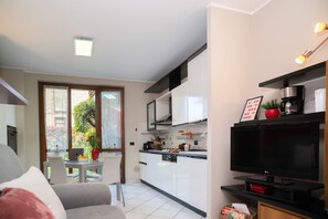 A modern kitchen with white cabinets, a black countertop, and a TV. There is a dining area with a table and chairs, and a view of greenery outside.