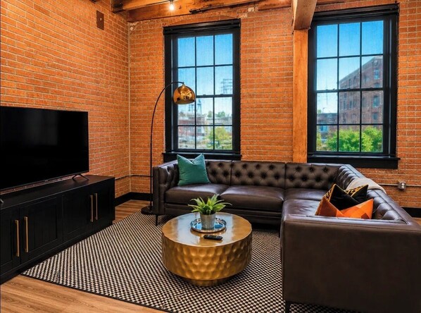 Ein Wohnzimmer mit Fernseher, Sofa und Couchtisch, mit Backsteinwänden und großen Fenstern.