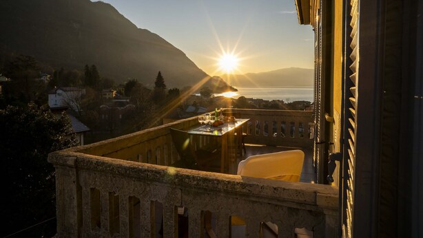 Een balkon met een eettafel, met uitzicht op een bergachtig landschap en een wateroppervlak tijdens zonsondergang.