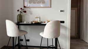 Uma área de refeições com uma mesa, duas cadeiras, um vaso com flores, uma cafeteira e uma obra de arte emoldurada na parede.