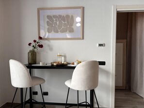 Ein Essbereich mit Tisch, zwei Stühlen, einer Blumenvase, einer Kaffeemaschine und einem gerahmten Kunstwerk an der Wand.