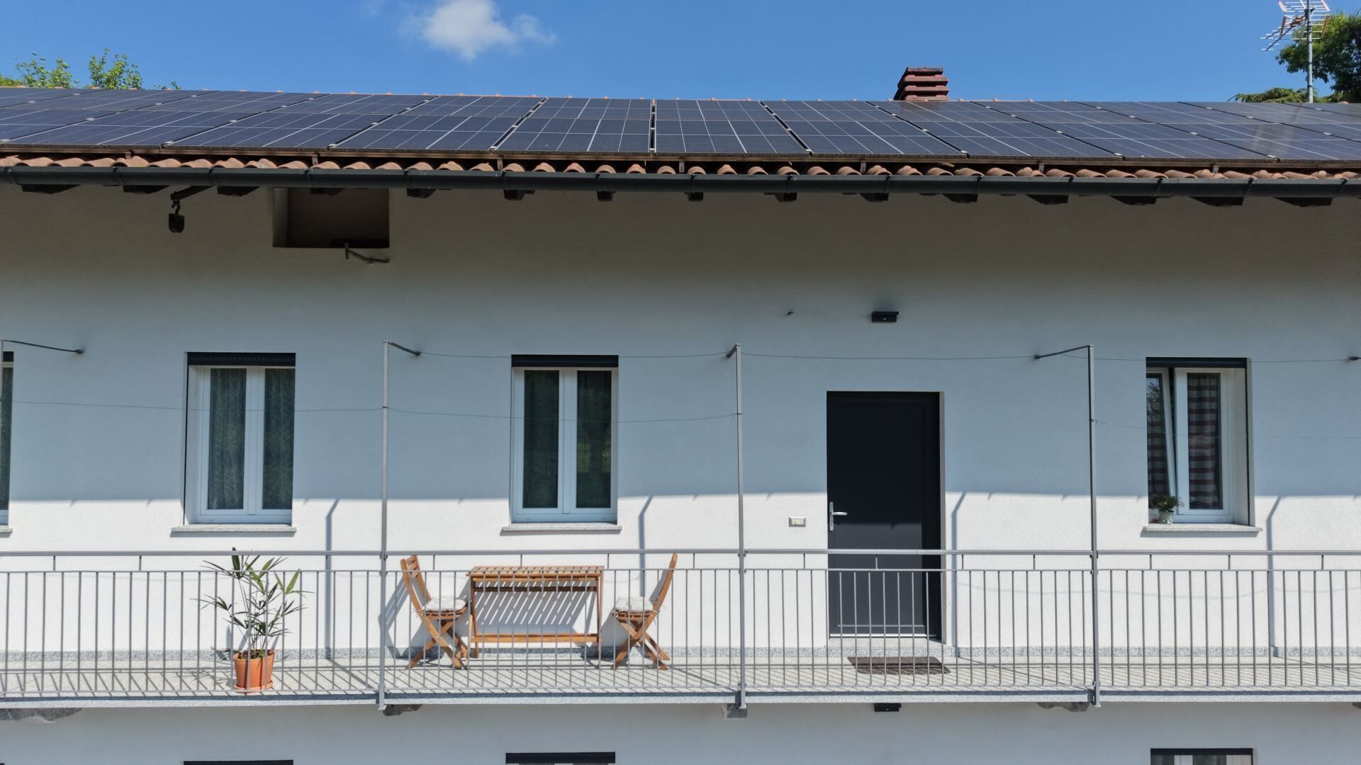 Ein Gebäude mit einem Ziegeldach, mehreren Fenstern, einer Tür und einem Balkon mit Stühlen und einer Topfpflanze.
