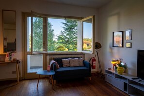 A living room with a sofa, a small table, a floor lamp, a TV, and a window with a view of trees and a house.