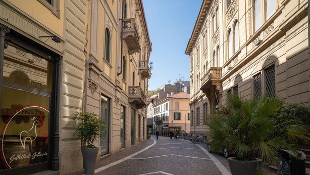 A narrow cobblestone street lined with historic buildings, featuring a shop with a rooster sign and potted plants. - Mazzini 1 - Smart Flat 2 bedrooms Garage (Gallarate)