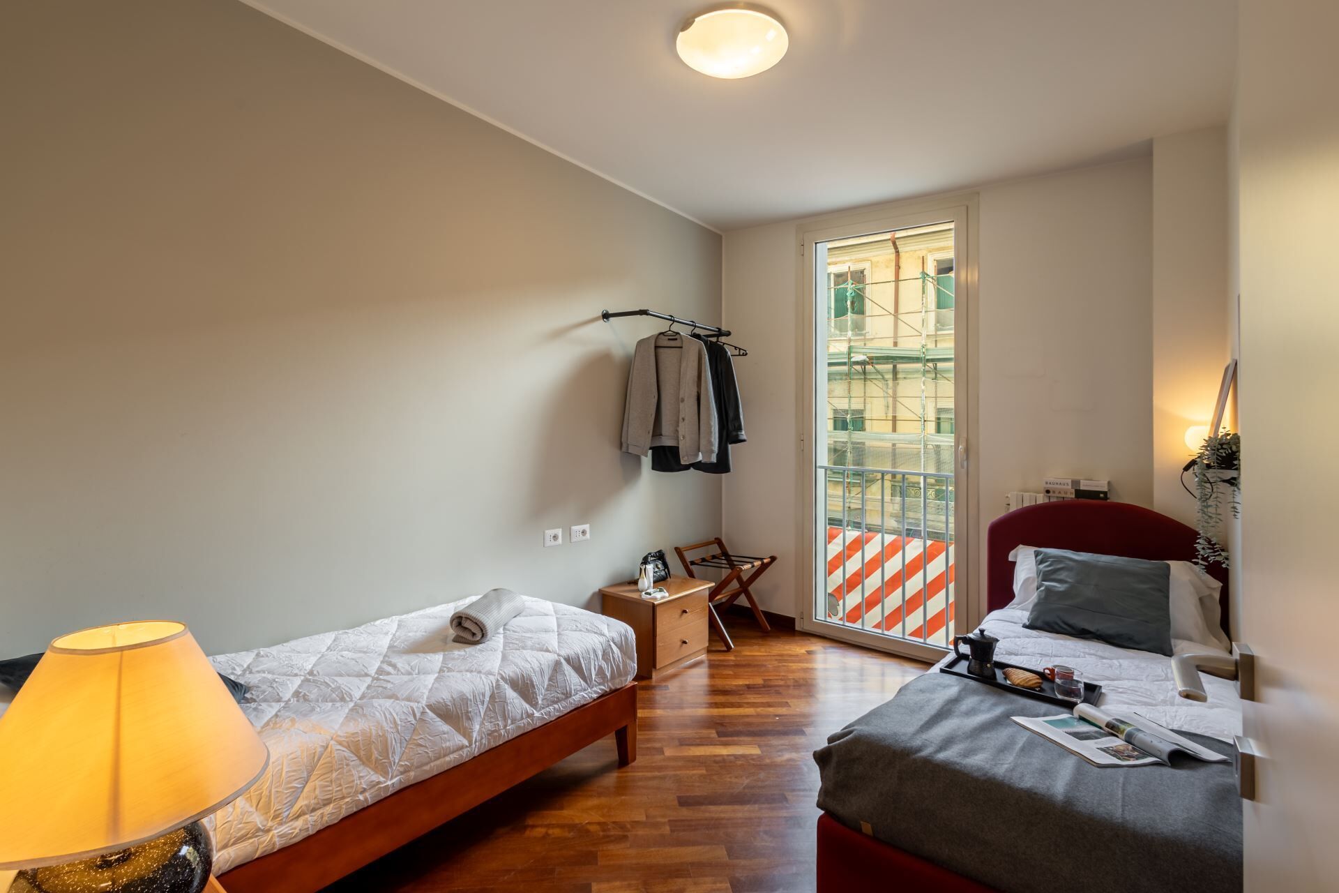 Una camera da letto con due letti, una finestra vista su un edificio, una lampada e un tavolino con un bollitore.