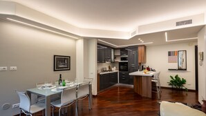 A modern kitchen with dark cabinets, a central island, and a dining area with a glass table and white chairs. - Mazzini 1 - Smart Flat 2 bedrooms Garage (Gallarate)