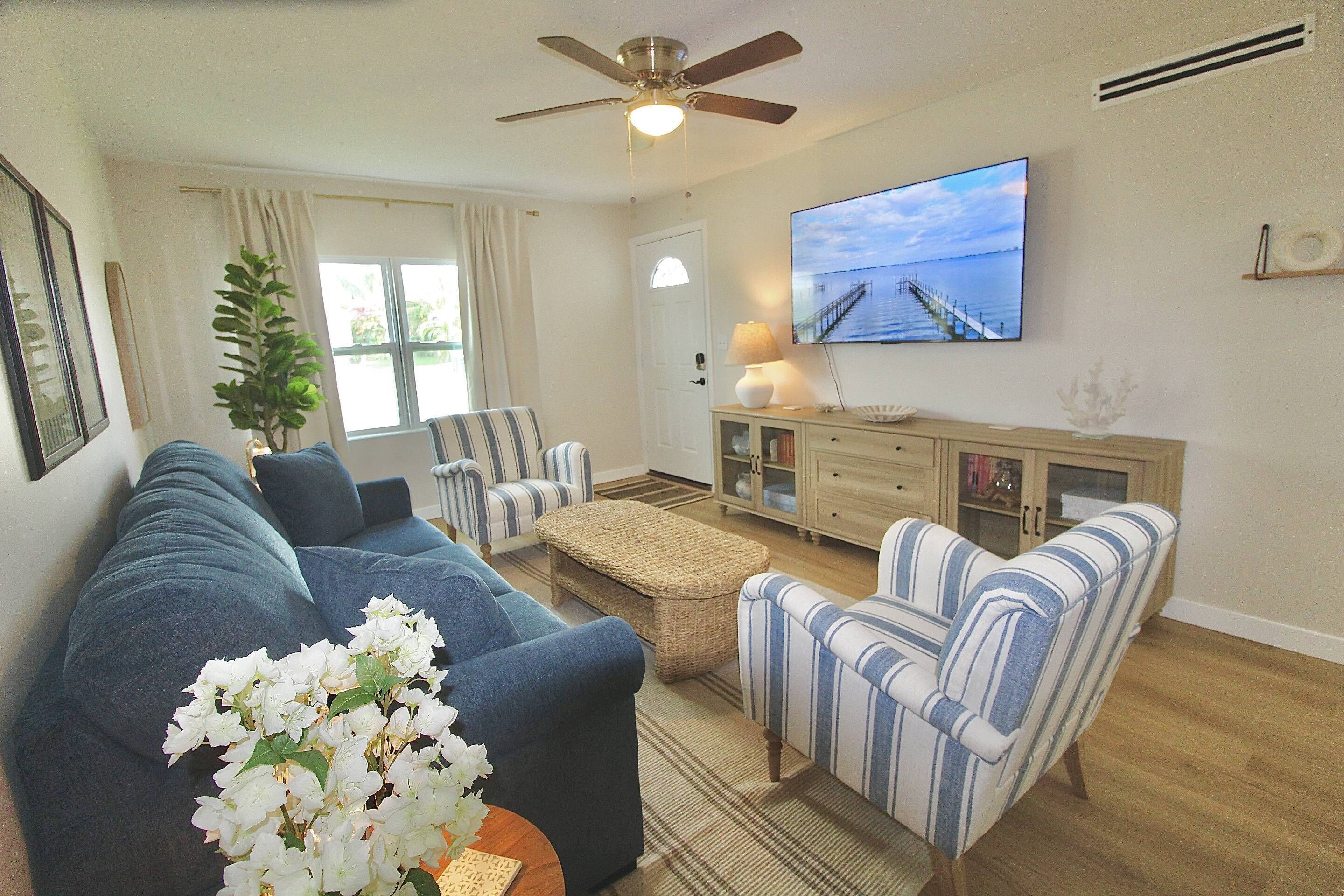 A living room with a sofa, armchairs, a television, a ceiling fan, and a window with curtains.