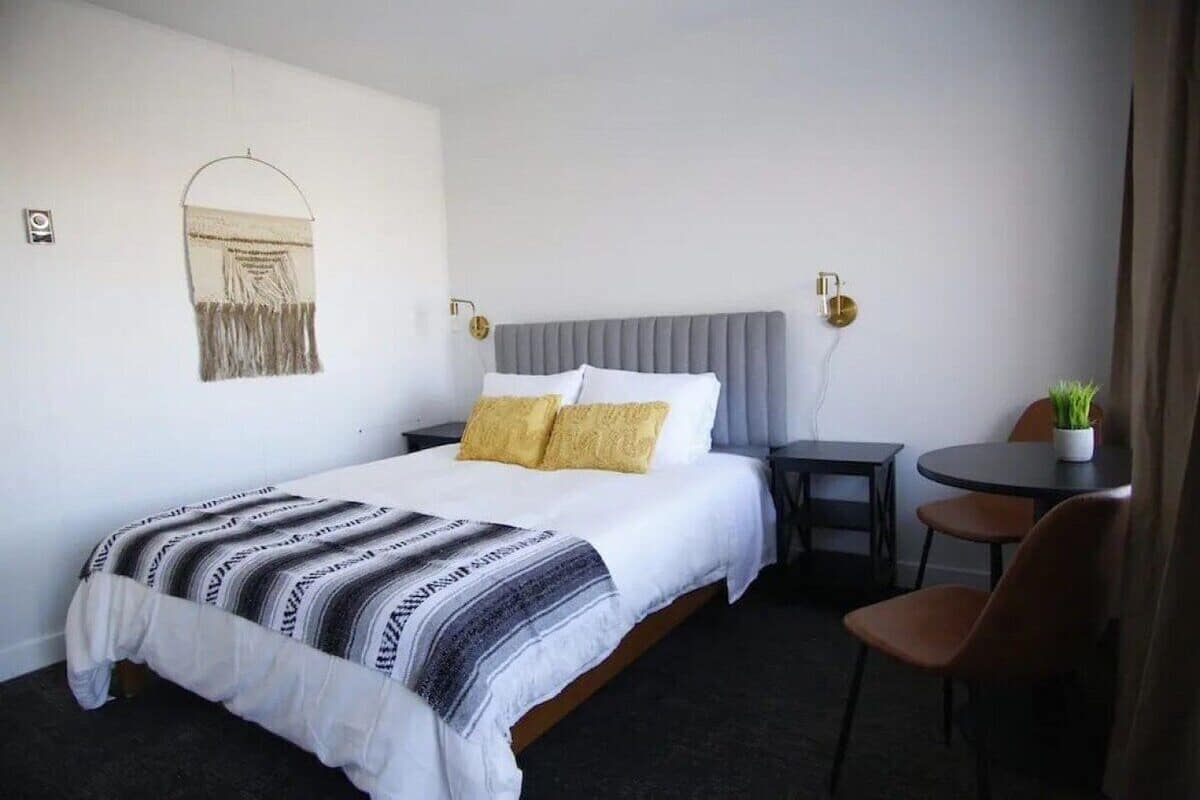 A hotel room with a bed, a bedside table, a chair, a small table with a plant, and a wall-mounted light fixture.