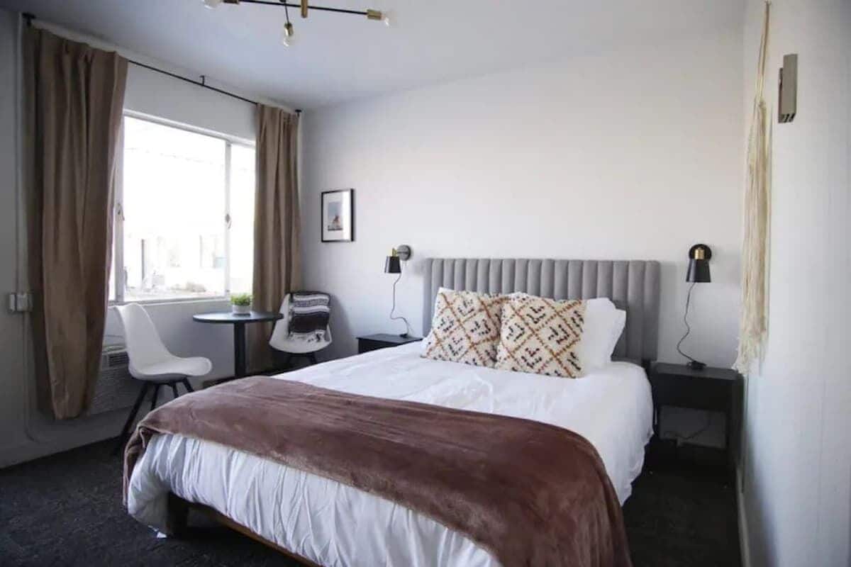 A hotel room with a bed, a bedside table, a chair, a small table, and a window with curtains.