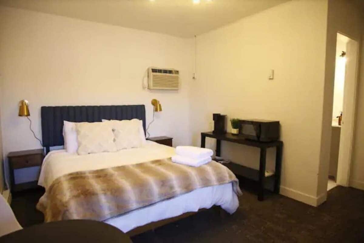 A hotel room with a bed, bedside tables, a desk, and an air conditioning unit.