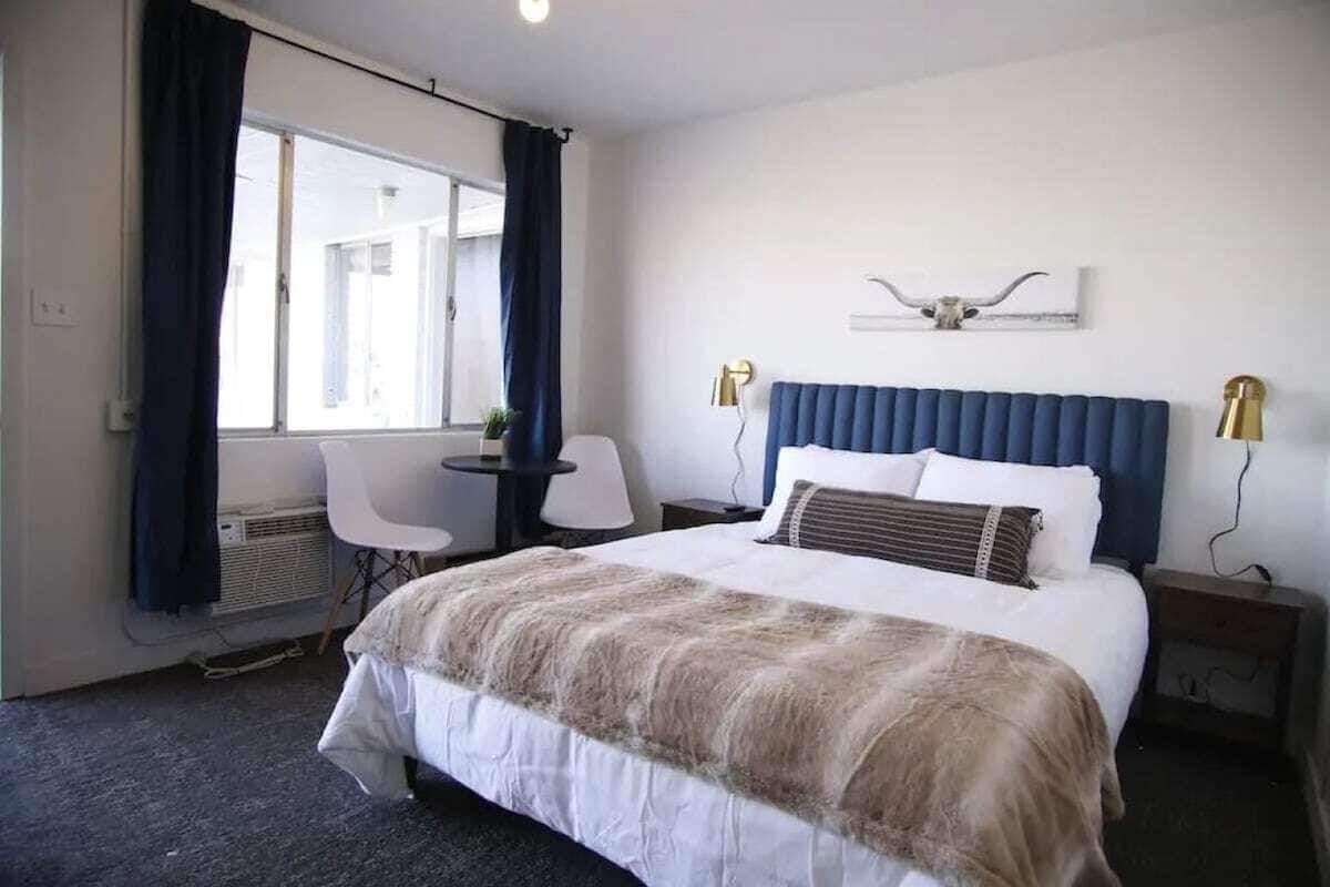 A hotel room with a large bed, a desk with a chair, a window with curtains, and a wall-mounted light fixture.