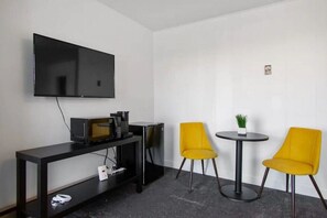 A small room with a flat-screen TV mounted on the wall, a black TV stand with a microwave, a small refrigerator, and two yellow chairs around a round table with a potted plant. - AEA - Fillmore UT (Fillmore)