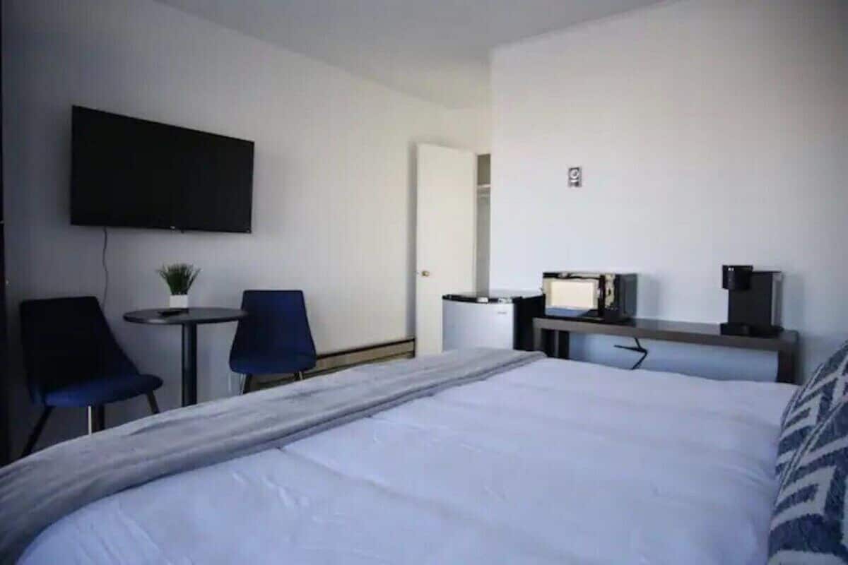 A hotel room with a bed, a desk with a computer, a TV, a chair, and a small table with a plant.