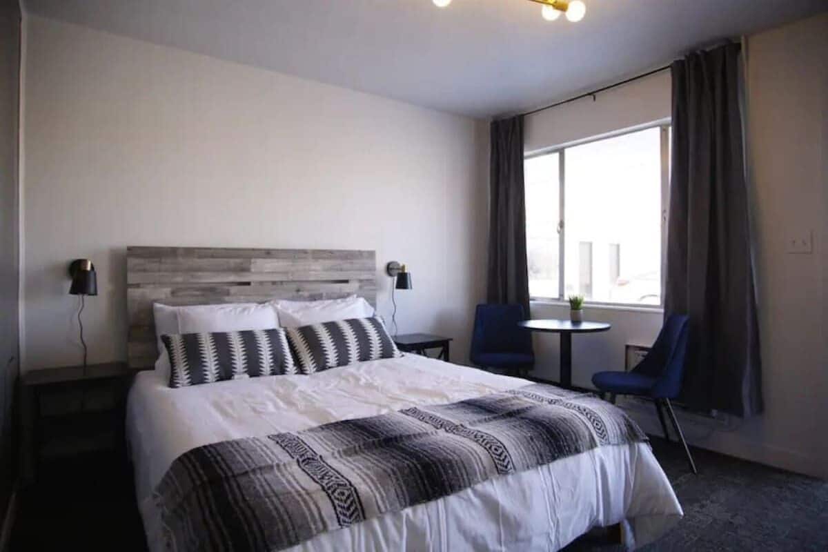 A hotel room with a large bed, a headboard, two bedside tables, a small round table, two chairs, and a large window with curtains.
